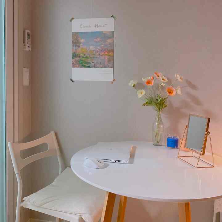 A small room in white and natural tones featuring a round table with chair, Claude Monet poster, and artificial poppy flowers in a simple setting