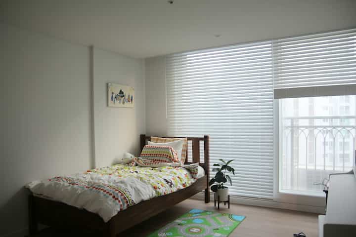 Bright and clean white and wood-toned kids' room featuring a single bed, white blinds, colorful bedding, and a small green plant creating a cozy atmosphere