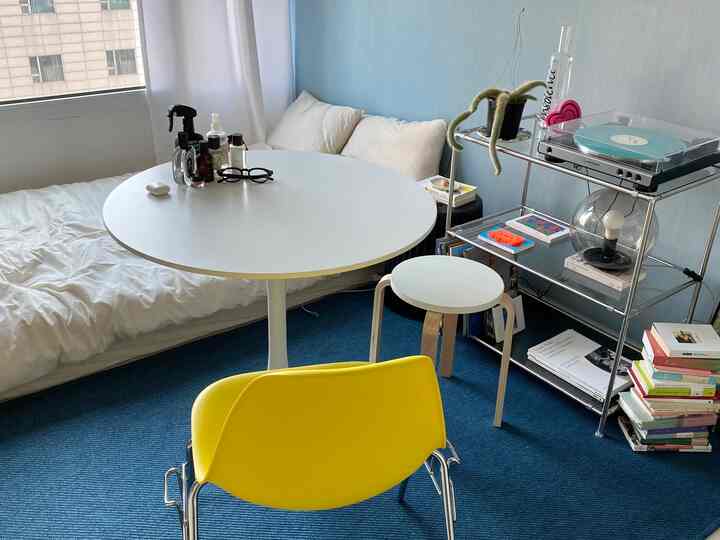 白と青のトーンのワンルーム、中央に丸いダイニングテーブルと黄色いチェア、モジュール棚の上にターンテーブルが配置された印象的な空間