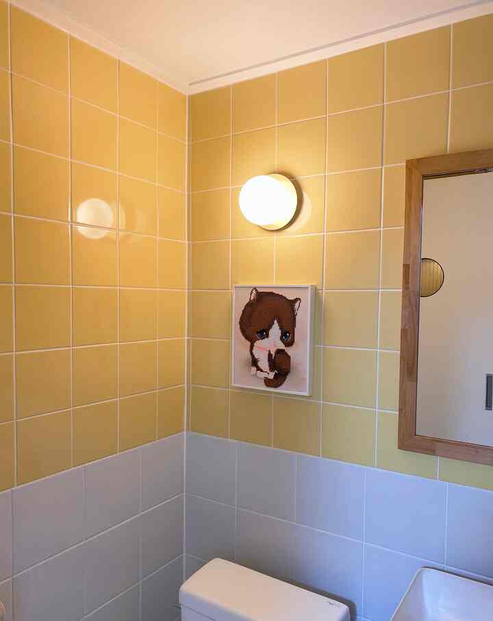 Bright clean bathroom with yellow and white tiled walls, toilet, wall-mounted light, and cat artwork