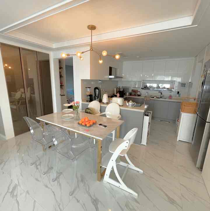 Dining room and kitchen in white and gray tones, featuring a marble dining table, transparent chairs, and kids' chairs in a modern, clean space