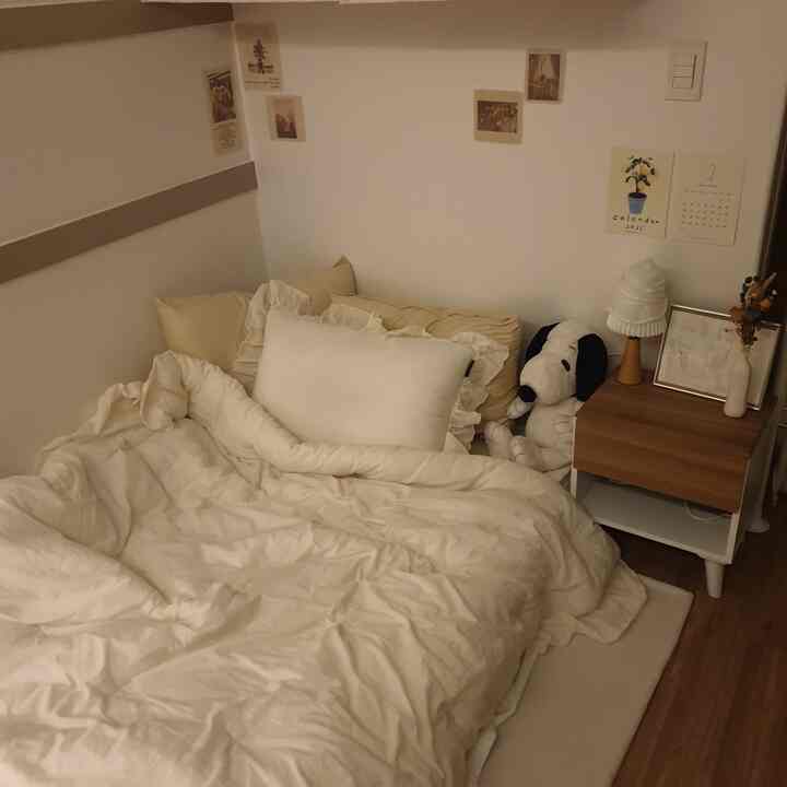 ベージュとアイボリーの色調の狭い寝室、居心地の良い寝具とナイトテーブル、壁に貼られた小さなポスターが温かみのある雰囲気を演出するインテリア