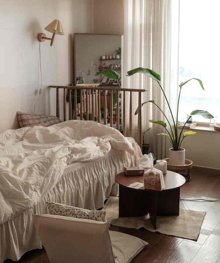 Beige-toned walls and wood-tone bed with white bedding in a studio bedroom featuring a small round table and large leafy plant with a natural atmosphere