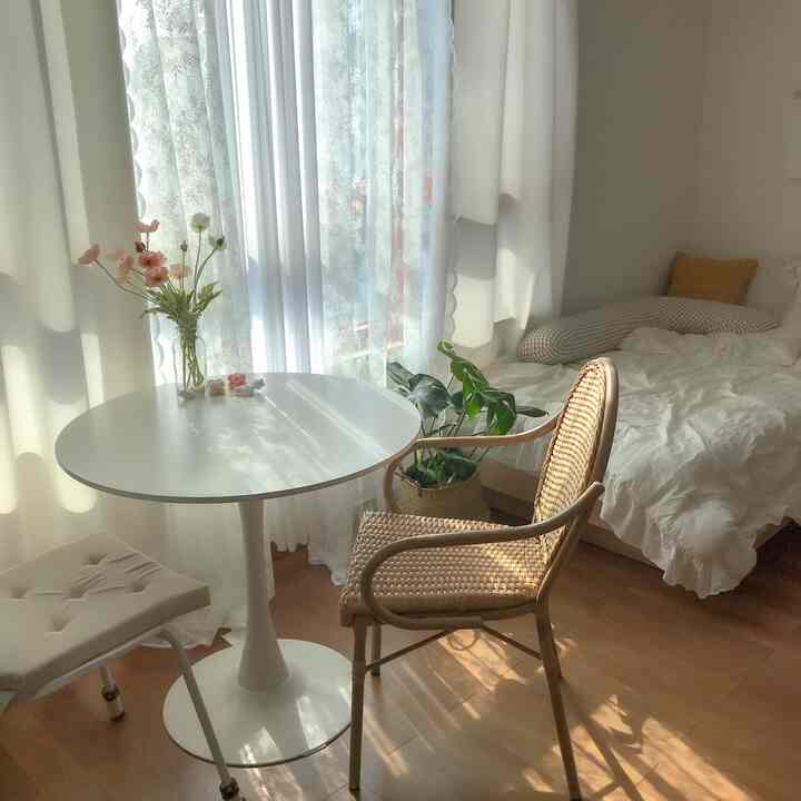 A soft wood tone and white bedroom featuring a rattan chair and round white dining table bathed in warm sunlight creating a natural cozy atmosphere