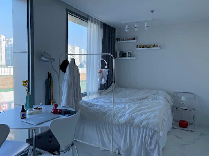 ホワイトトーンのワンルーム寝室空間、白いベッドとベッドカバー、透明な折りたたみチェア、二重カーテンが配置された清潔感のあるインテリア