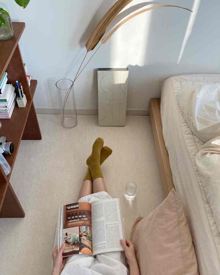 ベージュ調の寝室、ラグの上で本を読む姿と自然な木目調のベッドフレームが調和した雰囲気のあるインテリア