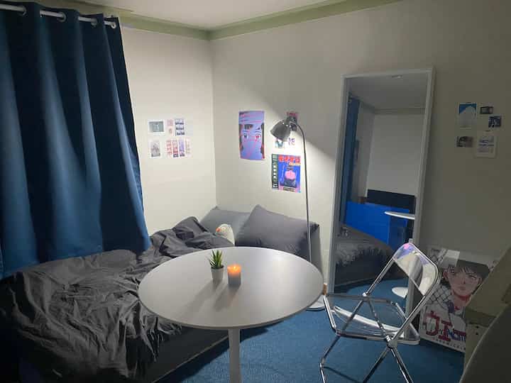 Compact bedroom in black and blue tones featuring a sofa bed, round table, and floor lamp creating a cozy atmosphere