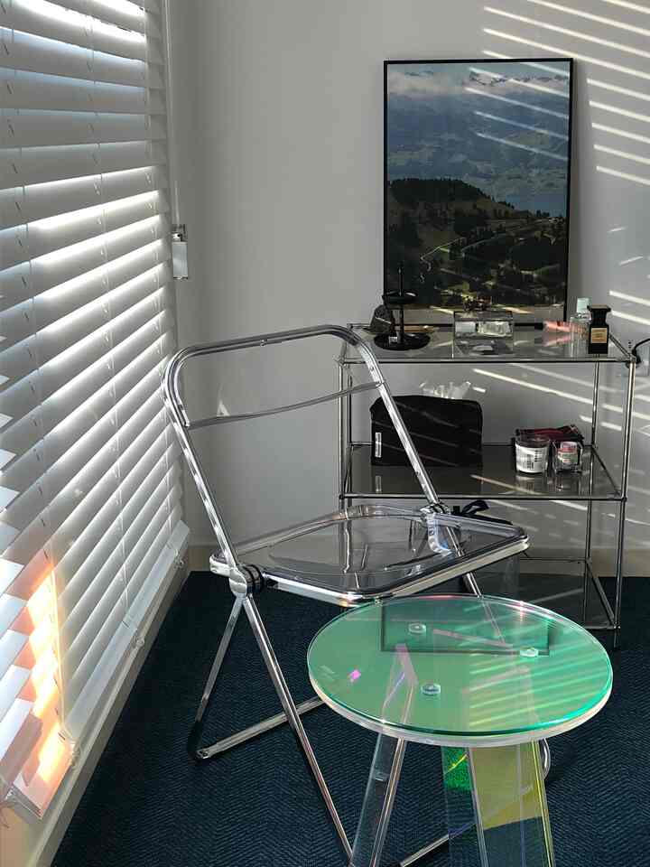 Modern interior featuring a transparent acrylic chair and holographic side table with white blinds softly filtering light