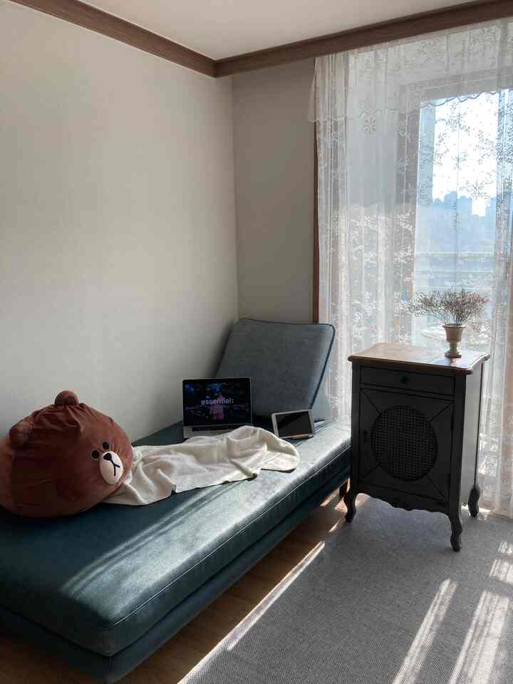 ブルーレザーのソファベッドと木目調のナイトテーブル、白いレースカーテンが配置された明るく居心地の良いリビング