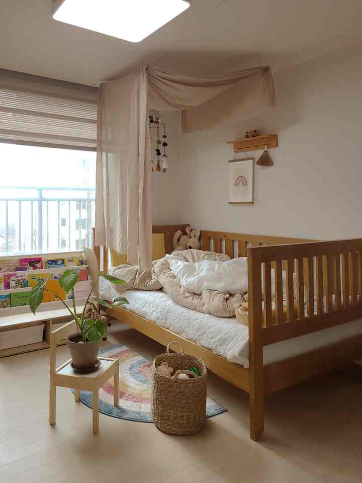 ナチュラルな色調の子供部屋、木製のベッドとベビーブックシェルフ、柔らかなベージュのカーテンと虹のポスターが配置された温かみのあるインテリア