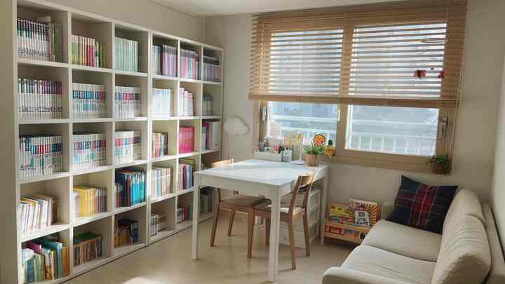 柔らかなナチュラルカラーの子供部屋、白い本棚とソファ、机、木製ブラインドが配置された雰囲気のあるインテリア