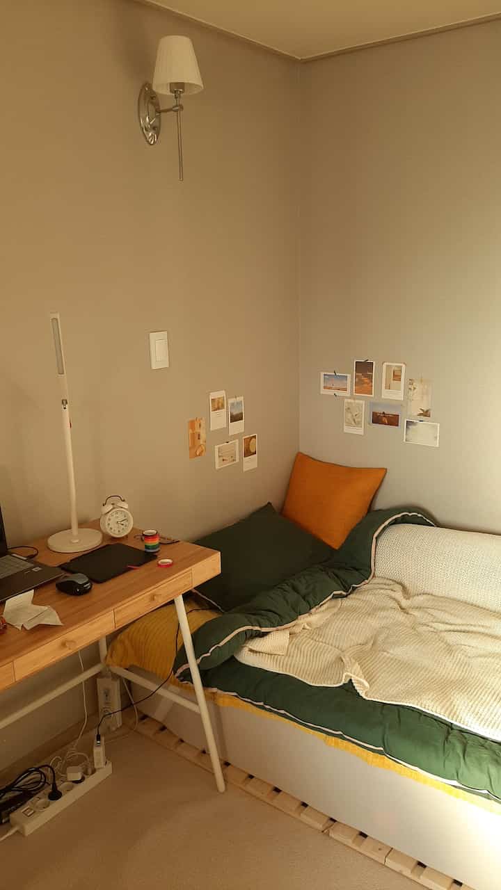 Natural-tone studio bedroom with wooden desk and cozy cushions, featuring aesthetic postcards on the wall for a warm atmosphere