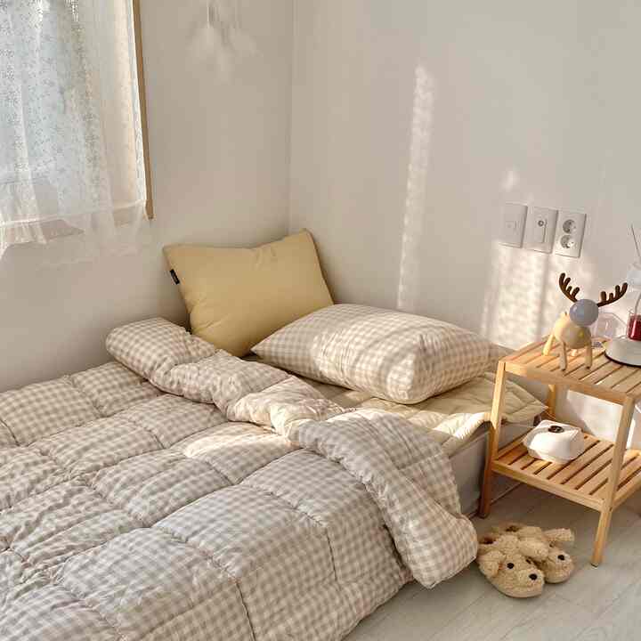 白い壁と木目調のナイトスタンドがある寝室、ベージュのチェック柄寝具と黄色い枕が温かみを演出する居心地の良い空間