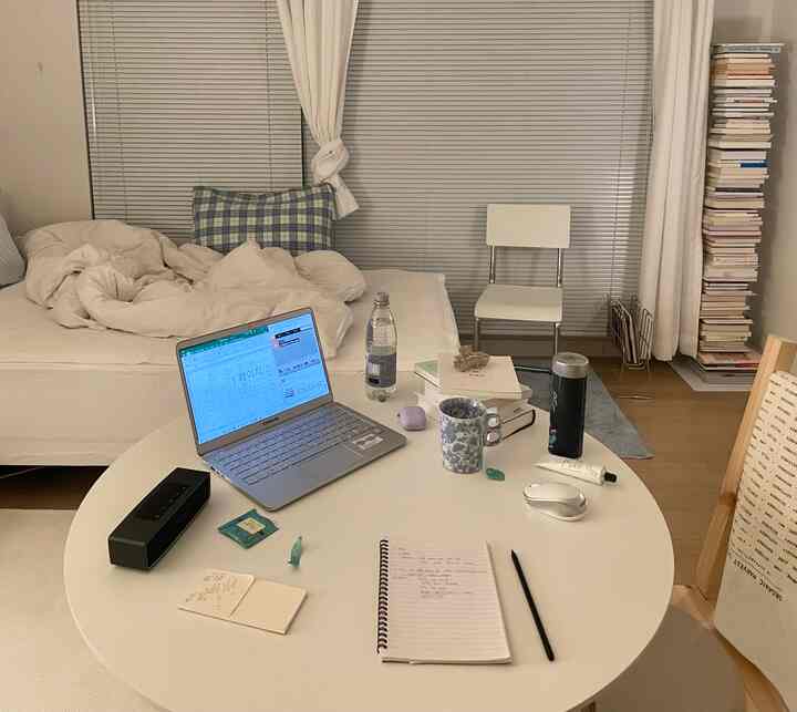 明るい白とベージュ調のワンルームリビング、ソファベッド、本棚、ブラインド、ノートパソコンとカップのあるデスクが配された落ち着いた機能的な空間