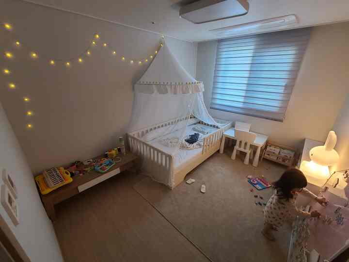 A cozy kids' room in soft white and beige tones featuring a canopy bed, LED mood lamp, toys, and a children's desk