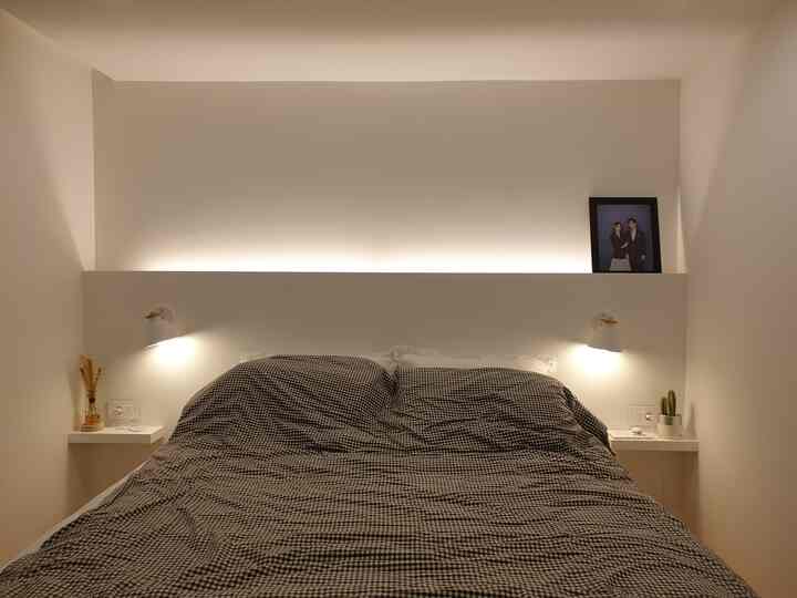 Cozy and simple two-person bedroom in white and wood tones, featuring a custom wooden headboard and warm indirect lighting