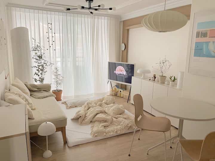 Bright white and beige natural-tone living room featuring a sofa, floor bed, and round dining table with cozy modern decor