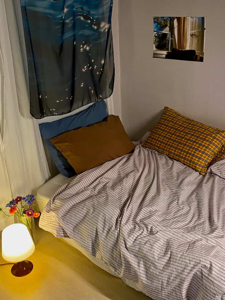 A white and wood-toned studio bedroom featuring minimal bedding and pillows, with a cozy table lamp creating warm ambiance