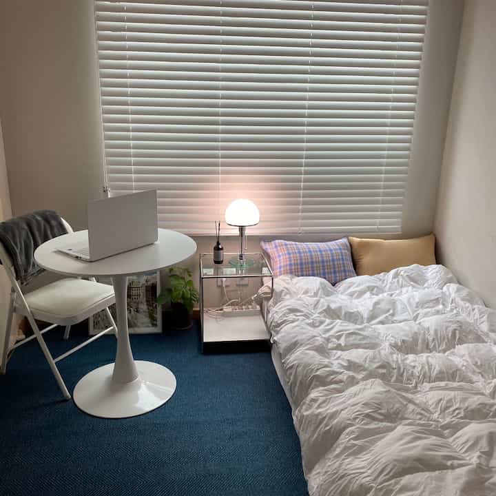 A compact bedroom in white and blue tones, featuring a bed, round table with laptop, and large white blinds creating a clean atmosphere