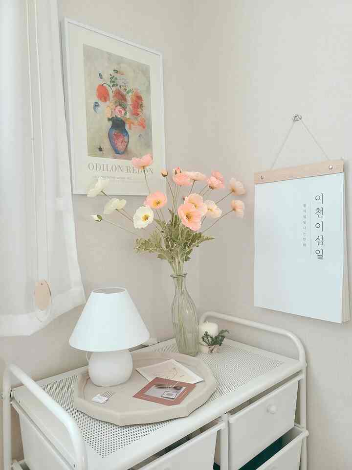 A cozy kids' room corner in white and beige tones featuring a tidy storage trolley, table lamp, and flower vase with floral wall art and calendar.