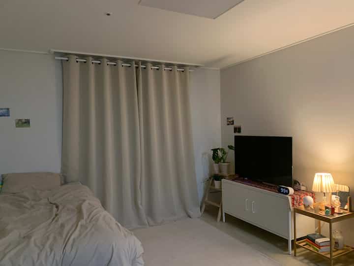 ベージュトーンのワンルーム寝室、大きなカーテンとベッド、テレビ台やナイトテーブルが配置された雰囲気のある空間