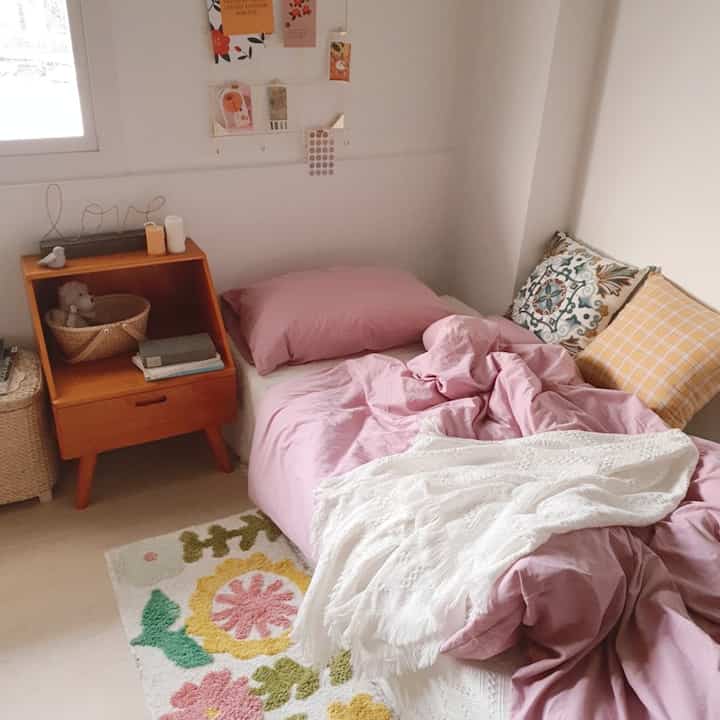 白い色調の壁とピンクの寝具が配置された4.5畳のワンルーム寝室、自然な木製家具が並ぶ温かみのあるインテリア