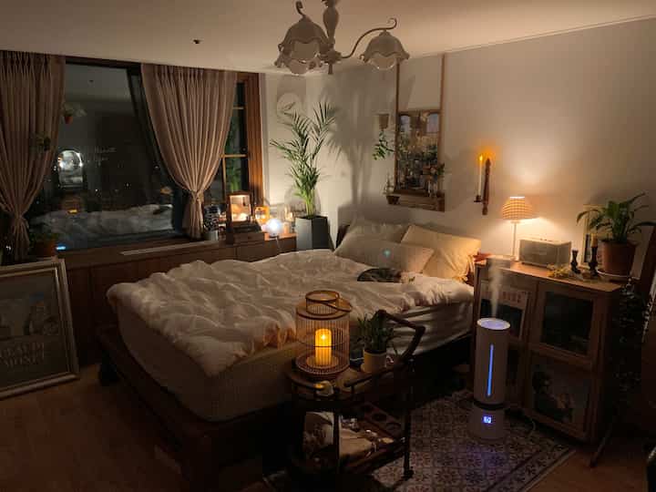 Cozy bedroom with warm wood tones and white bedding, featuring a humidifier and various plants creating a relaxing atmosphere