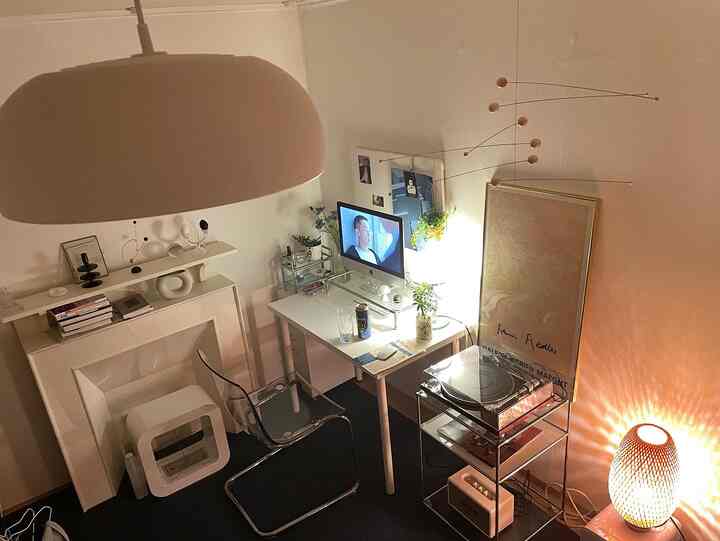 ホワイトトーンのコンパクトなホームオフィス、透明な椅子と白いデスク、モビールとポスターが配置され、柔らかな照明が居心地の良い雰囲気を演出。