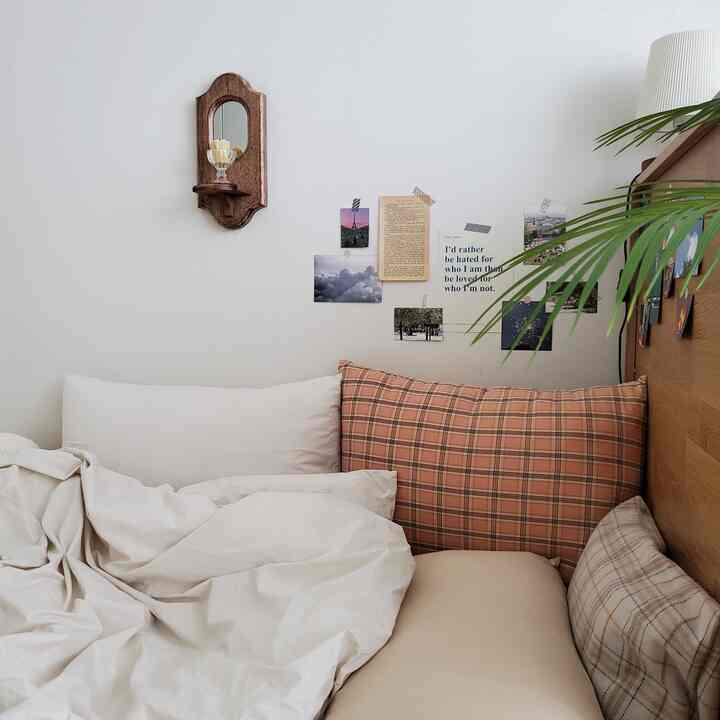 A cozy bedroom with beige and wood tones, featuring check-pattern pillows and sentimental postcards arranged on the wall