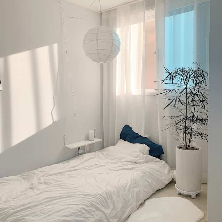 明るい白色の寝室、レゴリートのペンダントライトと白いカーテン、ネイビーの枕と大きな植物が配置されたシンプルで自然な雰囲気のインテリア