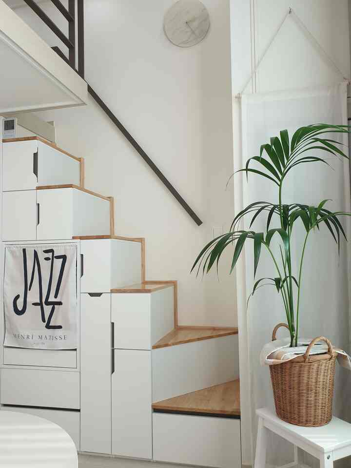 Natural modern staircase and storage area in white and wood tones, featuring a large plant and curtain with a cozy atmosphere
