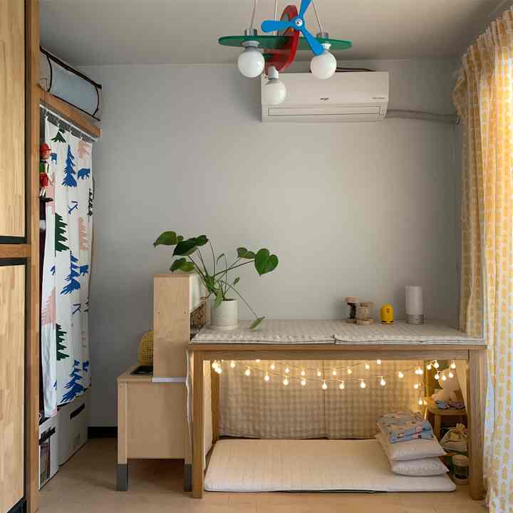 ナチュラルトーンの子供部屋、木製テーブルの下にカーテンとストリングライトで囲まれた居心地の良い秘密の空間があるインテリア