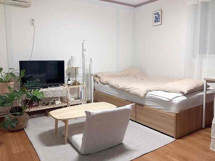 Natural and white-toned studio apartment bedroom featuring a bed, floor chair, rug, and plants creating a cozy resting area