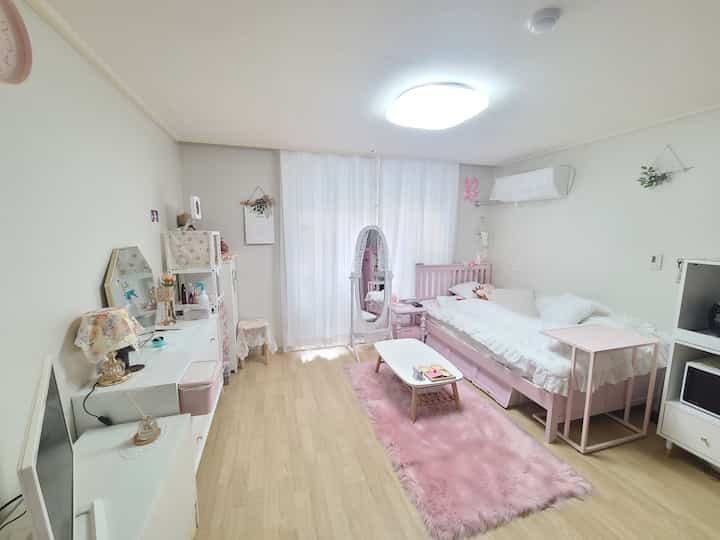 A cozy studio bedroom with bright white walls and ceiling, featuring a pink bed frame and a pink rug