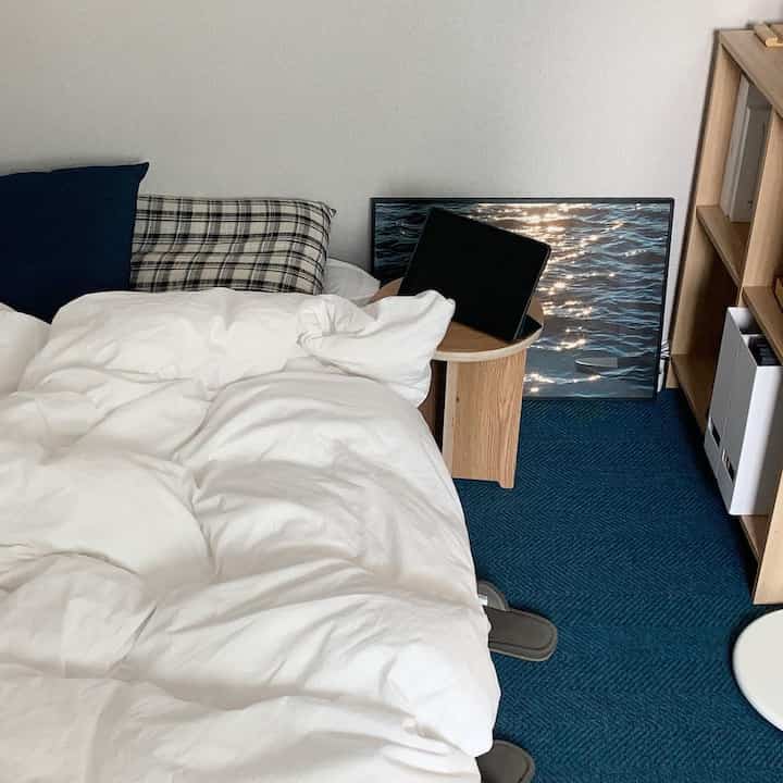 A bedroom space in white and navy tones featuring wooden shelves and a stool, with a minimalist and natural interior vibe