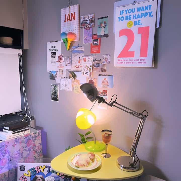 Light gray walled studio apartment featuring a yellow vintage desk lamp, postcards and posters on wall creating a cozy atmosphere