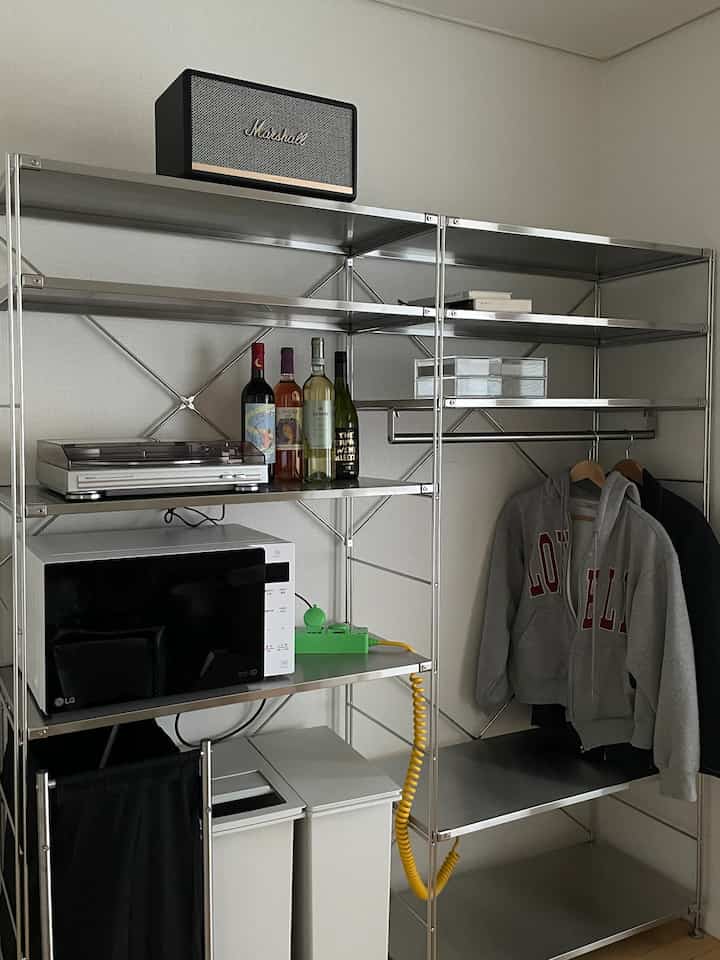 ステンレス製モジュラー棚、電子レンジ、ワインボトル、グレーのフーディが配置されたワンルームの印象的な空間