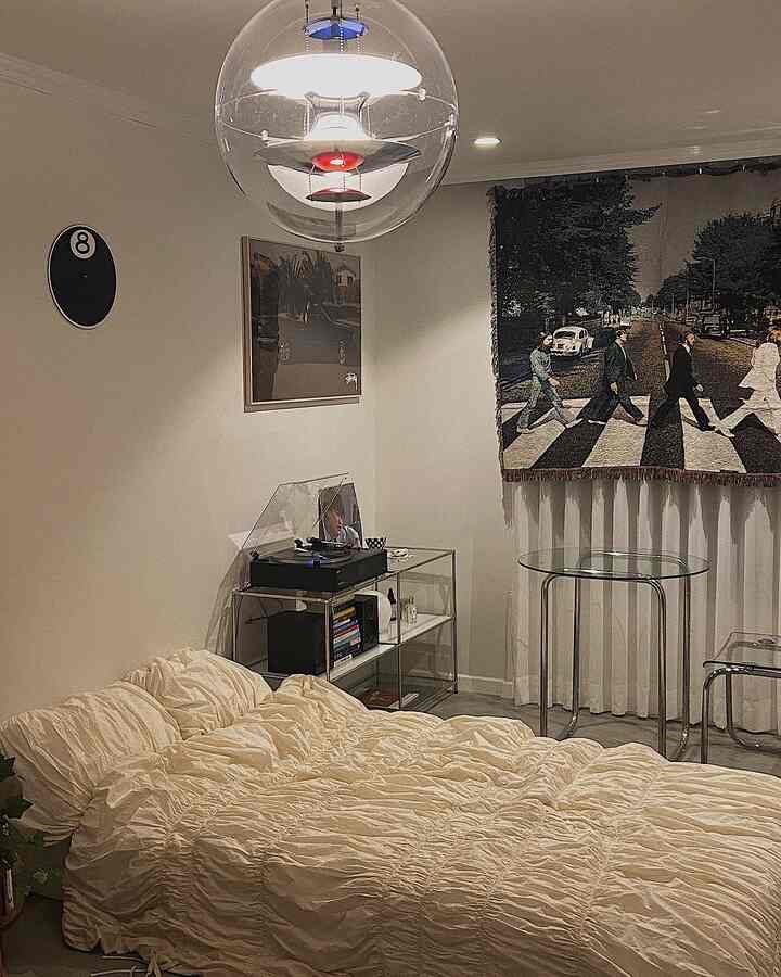 A bedroom with cream and beige tones featuring a bed at the center, transparent chair, glass table, and modern pendant lighting, creating a cozy modern atmosphere