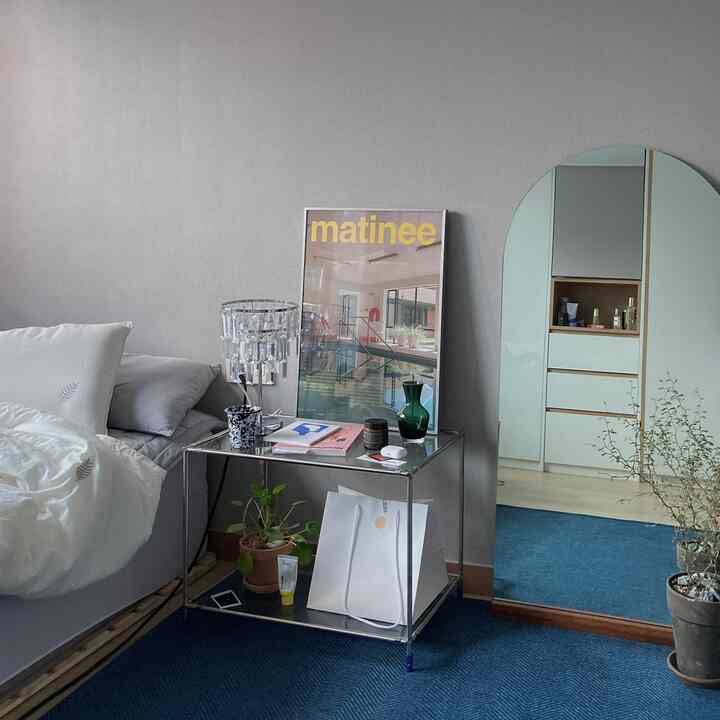 グレーの壁と青いカーペットが印象的な寝室、ガラスのナイトテーブルとテーブルランプ、ポスターが配置された雰囲気のある空間