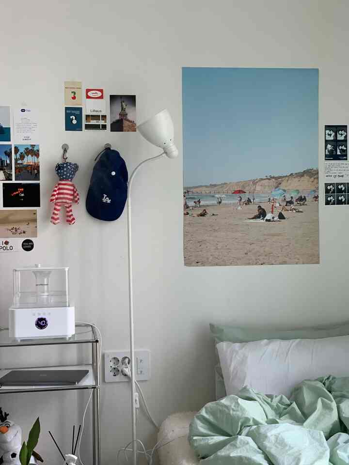 A small modern bedroom in white and mint tones featuring a bed, floor lamp, and various posters on the wall