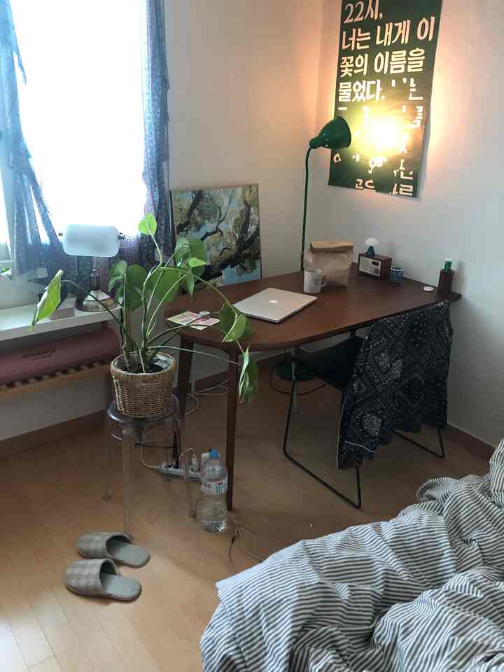 A white bedroom with wood tone floor and desk, featuring a laptop on the desk, floor lamp, and plant in a cozy home office setup
