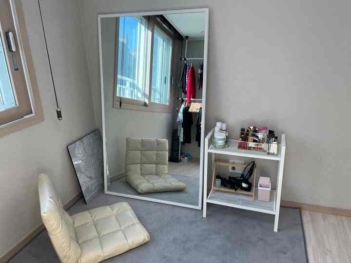 White-toned compact room featuring a large wall mirror, floor chair, and a cosmetics storage unit with a simple atmosphere