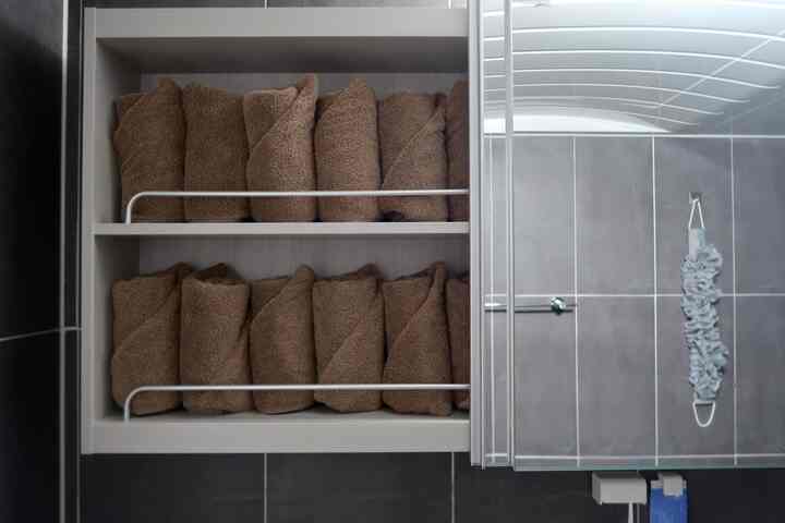 落ち着いたグレーのタイル壁と茶色のタオルが整然と並ぶスライド式収納キャビネットを備えたモダンなバスルーム