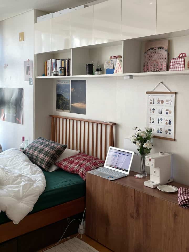 A compact bedroom featuring white and wood tones, with a bed, storage cabinets, and a laptop for remote work creating a cozy atmosphere