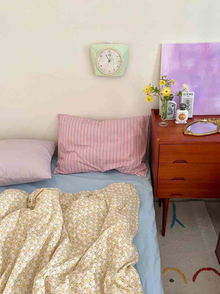 Soft pastel-toned bedroom featuring a yellow floral duvet, pink pillows, vintage wooden nightstand, and a mint clock on the wall creating a cozy atmosphere