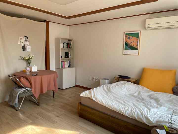 白い壁と木目調の床がある8畳のワンルーム寝室、小さなダイニングテーブルと本棚が配置された自然な雰囲気のある清潔な空間