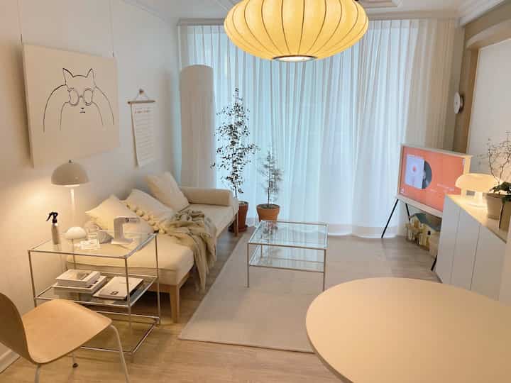 白とベージュのトーンのリビング、アイボリーソファと透明なガラステーブル、自然光と照明による明るく居心地の良い空間