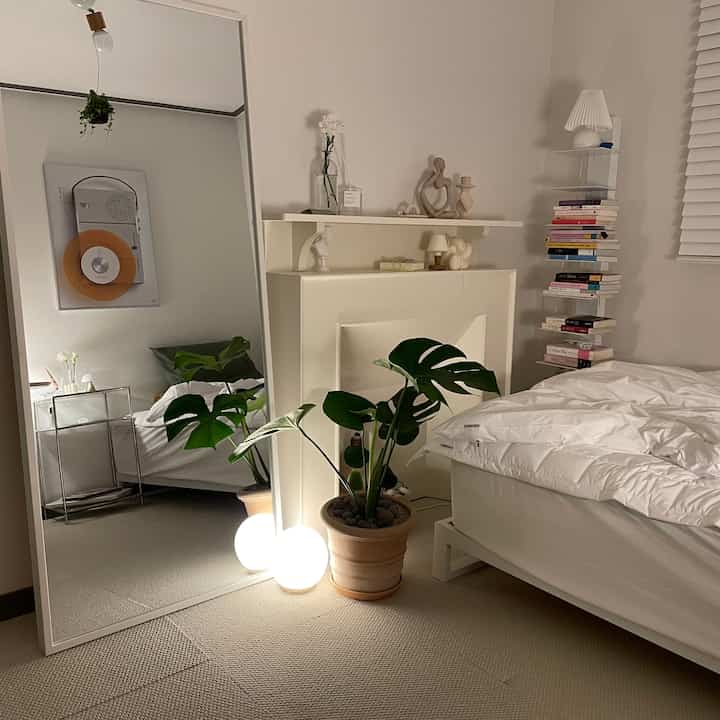 ホワイトとナチュラルカラーの寝室、大きな全身鏡とモンステラの植物が配置されたシンプルモダンで落ち着いた印象の空間