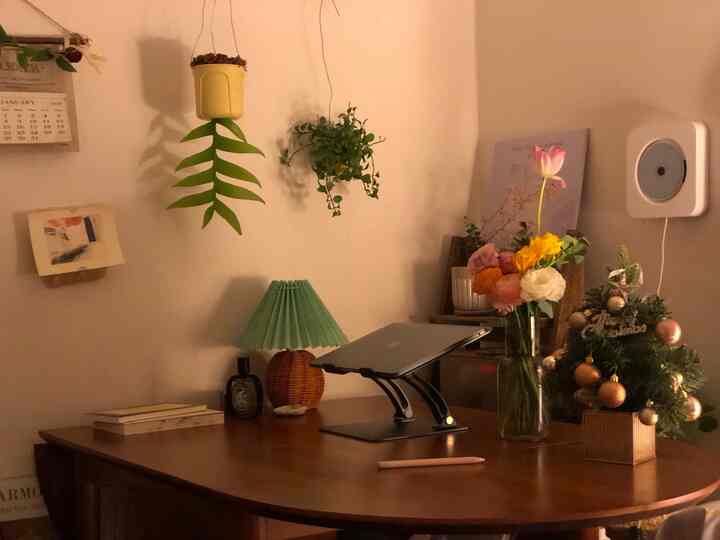 温かみのある木目調のリビング、デスクにノートパソコンと緑のテーブルランプ、花瓶と植物が配置された雰囲気のある空間