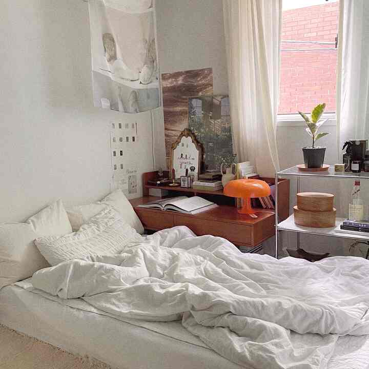 Bright white and natural wood-toned studio bedroom featuring bed, desk, and an orange table lamp in a cozy setting